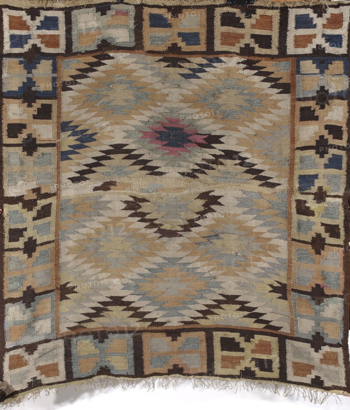 Kilim, Kilim, Podolia Ukraine origin; Poland cultural association. Decorative fabric for interior design, 1st half of the 19th century. Hand-stitched kilim with central field filled with rhombus motifs with stepped contours and slight overlapping in the center. Surrounded by a border of rectangle-like patterns with triangular notches on the longer sides. Geometric ornament. Colors: dirty gray, light blue and gray-blue, brown, ochre, brick red, with some navy blue and dark pink. Materials: fabrics. Height 141 cm; width 131 cm., A woven textile with a central field of zigzag and diamond motifs in muted beige, tan, light blue, pale gray, dark brown, and a small area of muted pink, surrounded by a geometric border of interlocking rectangular and plus-shaped motifs in beige, tan, blue, cream, and dark brown; the textile shows frayed edges and loose fringe at one end and variations in pile and wear across its surface.