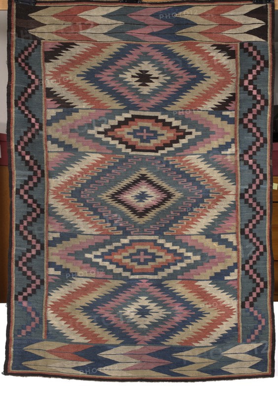Kilim, Kilim with vertical stripes in rhombus motifs that merge concentrically into hexagons, their contours formed by stepped lines. Borders: shorter sides wide with overlapping stepped-line motif shaped like the letter M; longer sides narrower with a zigzag-line motif. Geometric ornament; colors dark blue, white, brick red, and pink. Late 19th century, Poland. Fabrics. Height 178 cm, width 123 cm., A rectangular woven textile with a repeating geometric diamond and zigzag pattern in colors including blue, pink, red, cream, black, and beige; the design features a central vertical series of large diamond motifs surrounded by stepped chevrons and smaller angular shapes, flanked by vertical border bands with a stair-step motif, and top and bottom edges that show alternating elongated chevron forms; the overall surface shows tightly woven texture and symmetrical, angular motifs creating a layered, multicolored appearance.