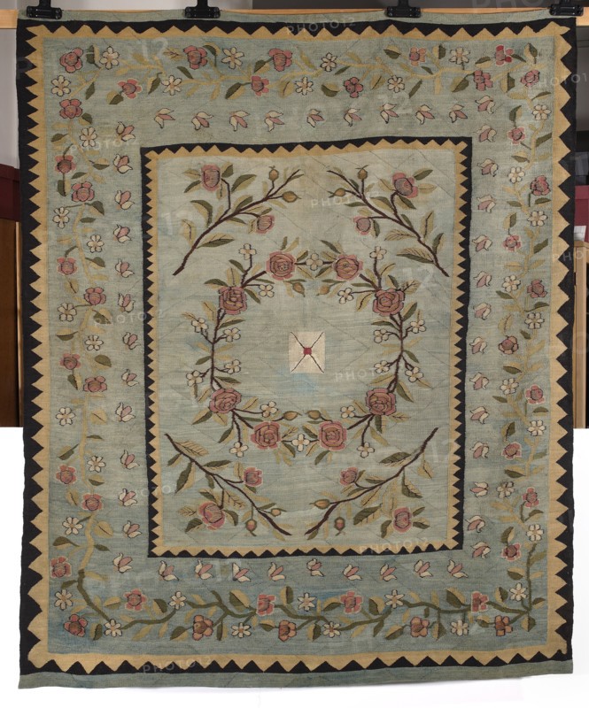 Kilim, kilim, court kilim, fabrics. In the center of the main field is a motif of a sealed envelope surrounded by a wreath woven with roses and small four-petal flowers and leaves. In the corners are motifs of twigs with flowers and rose buds. The kilim is surrounded by a wide border with a motif of a winding branch with bellflowers and six-petal flowers. This wide border is bordered on both sides by narrow zigzag trails. Colors: blue, white, pink, green, straw, ochre, and black. 1st half of the 19th century, Poland. Fabrics, height 206 cm, width 165 cm., A rectangular textile with a pale blue-green background features a central panel framed by a narrow dark border and a zigzag patterned band in dark and beige; inside the central panel there is a circular floral wreath of stylized roses and small flowers connected by curving branches and leaves in shades of pink, green, olive, brown, beige, and cream, with a small diamond-shaped motif at the center; surrounding the central panel is a w...