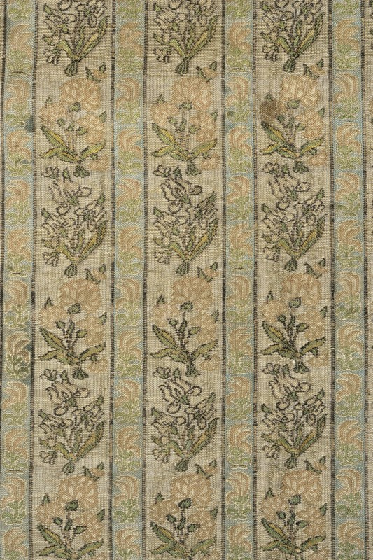 Fabric fragment. Gold-plated silk with a motif of irises and carnations in vertical stripes. Floral sprigs arranged in an alternating rhythm, turning first to the left irises and then to the right carnations. In narrower stripes, miniature stylized red flowers tulips?. Weave structure: compound weft-faced weave. Prominent wefts: gold thread with braiding, light and dark green, light and dark pink salmon, blue, white, black. Colors: gold background weft weave; patterns in green, pink-salmon, blue, black tied in weft weave. Main warp – dark yellow; main weft – cream. Dimensions of fragment: 8.0 x 4.3 cm; edge width: 0.5 cm. Selection width: 0.5 cm, fabric report: 4.3 x 8 cm. 18th century, Iran. Height 54 cm, width 28 cm., A textured vertical striped fabric with repeating floral motifs in a muted palette of beige, tan, cream, light green, pale blue, and dark olive; the design shows alternating narrow bands and wider panels, each wider panel containing stylized bouquets and leaves in beige...