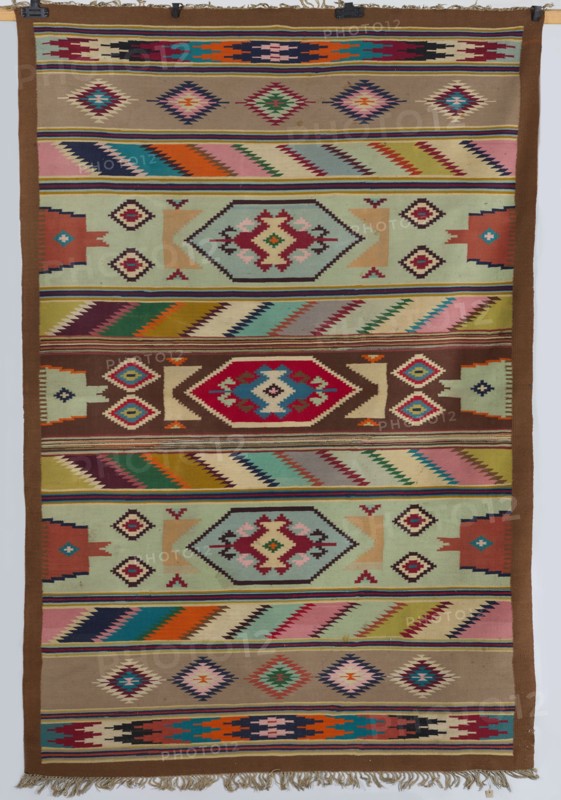 Kilim, The kilim is divided symmetrically into vertical stripes. The widest central stripe is decorated with an elongated hexagon containing other geometric motifs, including rhombuses and polygons. Adjacent stripes have diagonal serrated patterns; alternating stripes repeat the central hexagon motif in different colors, interspersed with more diagonal serrated bands. Further stripes feature rhombus motifs, and the two outermost stripes display intersecting rhombus motifs. No border. Colors: orange, white, brown, green, gray, brick red, and light celadon. 20th century. Materials: fabrics. Height 281 cm, width 191.5 cm. Poland., A woven textile with a repeating geometric pattern arranged in horizontal bands across its surface, featuring a palette that includes brown, tan, beige, cream, red, pink, orange, yellow, green, teal, blue, navy, and black; several bands show diamond and stepped motifs in contrasting colors, a few bands contain zigzag and chevron-like rows with alternating color ...