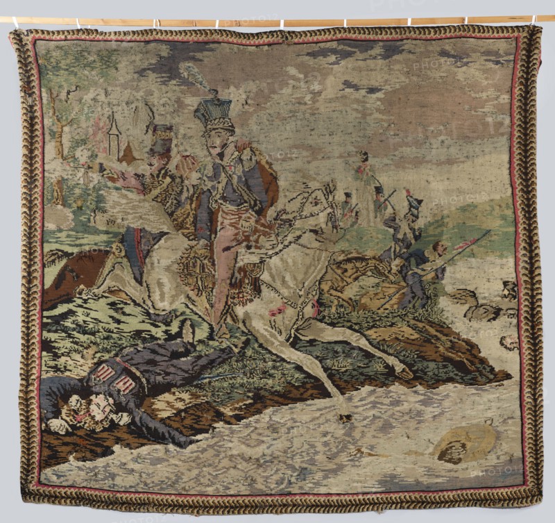 Kilim depicting the death of Prince Józef Poniatowski, after Horace Vernet 1789–1863 and an unknown author, early 19th century 1st half, wool fabric tapestry, height 213 cm, width 220 cm. Scene: on the right a river, center Prince Józef and his soldiers, upper left the outline of a city; colors: brown, cream, red, blue., A woven scene shows a group of figures and horses in an outdoor setting rendered in muted colors including beige, tan, brown, green, blue, pink, and red. A central figure in a decorative uniform with a tall hat rides a pale horse that is stepping forward, while nearby another rider on a darker horse gestures outward. Several other figures in uniform appear in the middle and background, some carrying long poles or rifles. In the foreground, a fallen figure lies on the ground wearing a dark coat with lighter trim. The landscape includes grassy mounds, rocky ground, and a body of water depicted with pale and grayish tones, and a round object floats near the water’s edge. ...