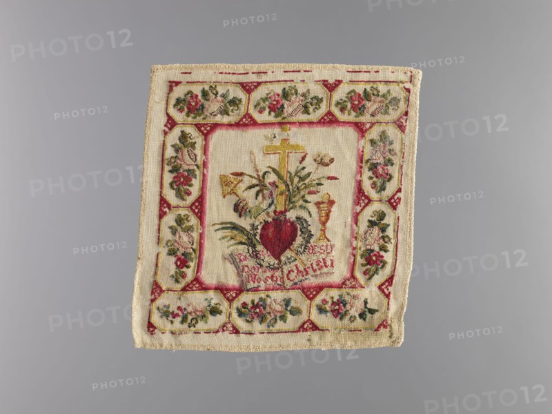 Pall, origin unknown, church parament featuring crosses, chalices, and plant ornaments; sacred art for churches and chapels furnishings; liturgical parament, 1840s; height 17 cm, width 17 cm, A square textile with a central embroidered design surrounded by a decorative border is visible; the fabric is a light beige or cream color with embroidered areas in red, pink, green, yellow, gold, and dark brown. The central panel shows a heart-shaped motif in deep red with surrounding green leaves and stems, a gold-colored cross above it, and a chalice-shaped form in gold nearby, all stitched on the cream background. The surrounding border consists of a repeating pattern of floral medallions and geometric elements in red, pink, green, and gold on the same cream fabric, with some darker brown and black accents in the embroidery. The textile edges are finished with a woven trim in beige, and the overall piece shows slight signs of wear and irregularity along the edges.