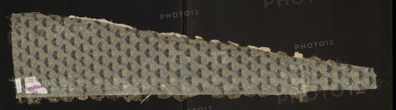Fragment of a garment, fabrics with a preserved report, silk fabrics, floral patterns, fragments of a larger textile object. Design: small silver twigs form a net with meshes in the form of palmettes inside which white roses are placed; blue-silver stems. Report: 3.7 x 2.7. Fabric density in 1 square cm: 31 white warps, 31 blue wefts. 17th century, Iran, fabrics. Height 76 cm, width 17.6 cm min. 4.3 cm., A long, irregularly triangular fabric fragment laid flat against a dark background, showing a repeating small geometric motif that alternates between rounded and fan-like shapes; the fabric surface appears worn with some frayed edges and loose threads along the top and bottom borders, and a section of fringe remnants along one long edge. The visible colors include muted blue-gray as the dominant ground, darker navy or deep blue within the motifs, and a faded beige or tan accenting other parts of the pattern and the frayed fibers, with small patches of off-white where the material is mo...