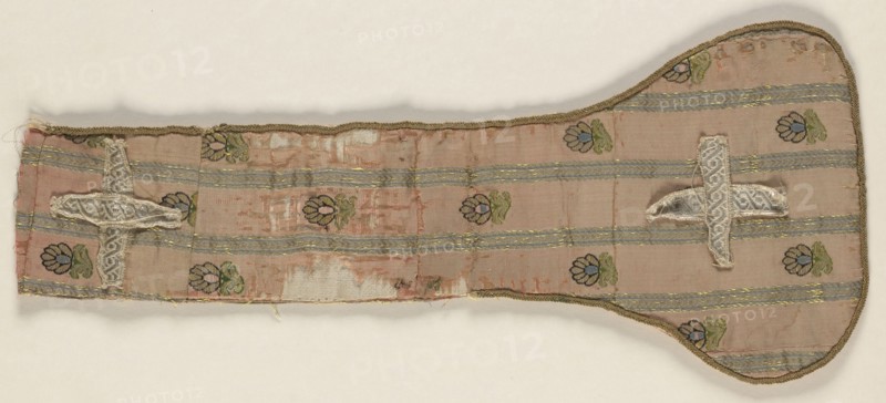 Fragment of a maniple made of crimson silk fabric with a striped and fine-thread motif. Salmon background, gold stripes and throws with black, green, and blue accents. The preserved fragment contains one ending pallette with slightly rounded edges trimmed throughout with a round wire. An isosceles cross made of silver braid is sewn onto the pallette. Identical fabric preserved in two fragments cut into stole pallettes. Fabric used for sewing zupans and kontusz lapels, probably from Grodno. Pattern motifs: small flower sprigs between gold stripes. Colors: pale red grosgrain background; trim: gold metal thread; brocades: pale blue, black, light green, gold, yellow. 2nd half of the 18th century, Poland. Liturgical vestment; length 43 cm, width 18 cm., A long, narrow fabric piece with a rounded wider end, primarily pink with a faded, worn appearance, showing a pattern of horizontal pale blue-gray stripes and small floral motifs in green, dark blue, and lighter blue. The fabric has metallic...