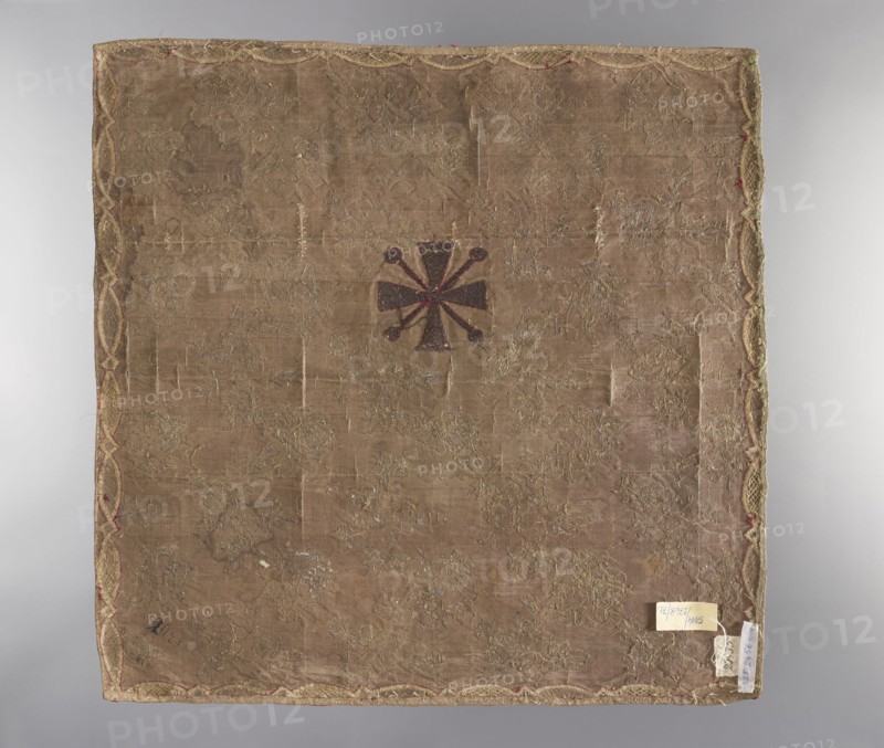 Velum; unknown creator; ; church paraments; plant ornaments; sacred art; churches and chapels furnishings; 18th century; France; fabric; liturgical parament; height 50 cm; width 50 cm, A square piece of fabric with a worn beige-brown ground and a decorative scalloped border in golden and reddish tones, showing a central dark brown emblem with radiating petal-like shapes and small round ends; the surface has scattered areas of metallic embroidery in faded gold and silver hues and visible patches of wear and thinning; the edges show a few small tags and remnants attached, and the overall palette includes beige, brown, gold, silver, and hints of red.