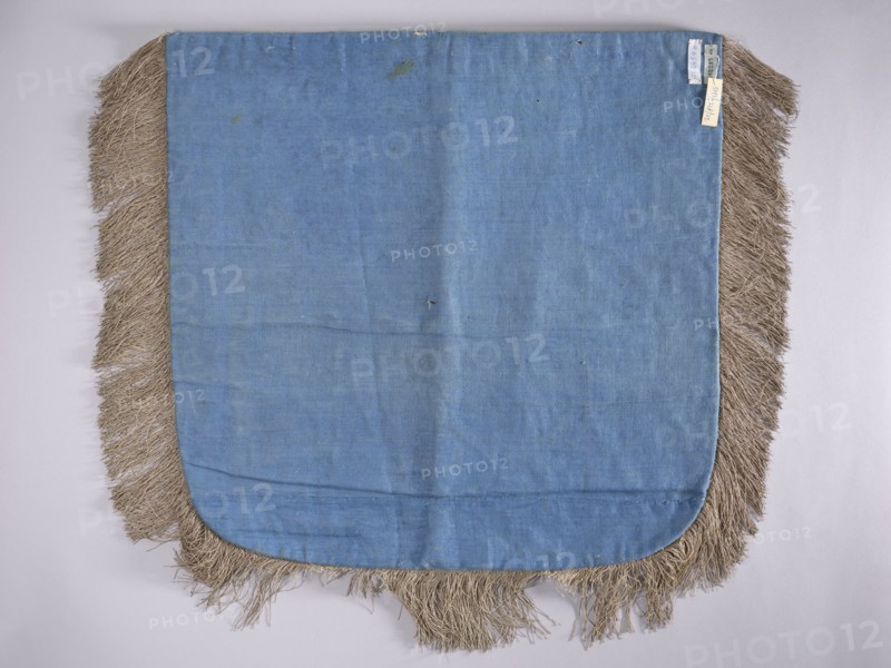 A hood from a church cope. The cope hood is made of pale green velvet. In the center, two embroidered palmettes run parallel to each other, surrounded by plant-braided motifs, with a finer ornament embroidered with a single thread closer to the edges. The hood is trimmed with silver braid: wider at the top, narrow around the remaining edges. The edges are fringed. 18th century, Poland. Liturgical vestment. Height 53 cm, width 52 cm., A rectangular fabric panel with rounded lower corners, mostly light blue in color, bordered on three sides by a fringe made of coarse beige fibers; the blue fabric shows several faint creases and a few small darker spots, and the fringe appears uneven and slightly matted in places against a pale neutral background.