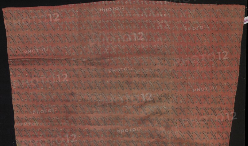 Fragment of fabric from a garment, Silk Factory Company, fabrics, silk, 2nd half 18th century. Decoration: small leafy twigs and two buds repeated in rows, alternating. Made on a wide loom over 78 cm; colors: crimson background with a rep weave; brocades in navy blue and ochre-silver. Pattern repeat: 4 x 2.5. Same fabric — PIECE 247. Place of origin: Grodno, Lithuania Grand Duchy of Lithuania; 1569–1795; Poland. Fabric, height 113 cm, width 39 cm., A rectangular piece of fabric with a muted reddish-pink background laid against a dark background, covered in a repeating diagonal vine-like pattern in darker grayish-blue and pale beige tones, showing subtle horizontal creases and slight variations in the red color with some areas appearing more faded or darker, and narrow black margins visible along the left and right edges.