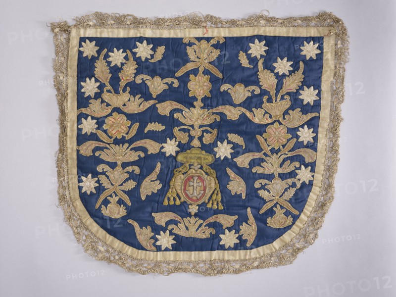 A hood from a church cope. The hood of the liturgical cope is made of navy blue silk with appliquéd decoration of stylized flowers, leaves, and stars. In the lower center, beneath the bishop's hat, is a cartouche with the Odrowaz or Dolega coat of arms. Predominant colors are gold, silver, green, and brick red. The hood is trimmed with contemporary metal lace braid. The appliqué was transferred to a new fabric. 18th century, Poland. Fabrics. Height 58 cm, width 61 cm., An embroidered textile panel with a deep blue fabric background bordered by a pale gold trim and an outer fringe of metallic threads, featuring an array of ornamental floral and foliate motifs in shades of gold and muted pink set against the blue, a group of star-shaped floral forms in pale gold scattered across the surface, and a central decorative emblem with gold elements and touches of muted pink and red surrounded by symmetrical scrolling leaf and petal designs.