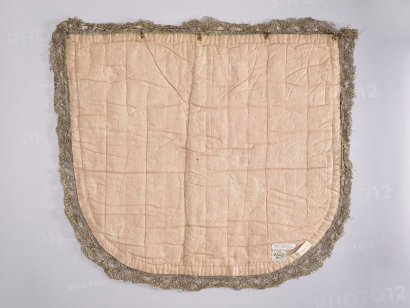 A hood from a church cope. The hood of the liturgical cope is made of navy blue silk with appliquéd decoration of stylized flowers, leaves, and stars. In the lower center, under the bishop's hat, is a cartouche with the Odrowaz Dolega coat of arms. Predominant colors: gold, silver, green, and brick red. Trimmed with contemporary metal lace braid. The appliqué was transferred to a new fabric. 18th century, Poland. Materials: fabrics. Height 58 cm, width 61 cm., A rectangular fabric panel with a rounded bottom edge is visible, mostly pale pink in color and showing many creases and stitched quilt-like lines; it is edged with a decorative fringe in a metallic grayish-gold tone that forms an irregular border around the fabric.