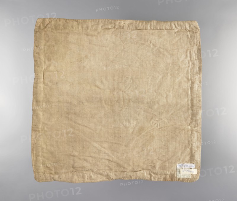 Velum. Velum made of blue silk fabric. In the center is an applied radiant medallion with the IHS monogram with a cross and a burning heart. In the corners and in the center of each side are traces of torn-out applied flower sprigs. Trimmed with wide silver braid. Linen lining. 18th century, Poland. Liturgical vestment. Height 45 cm, width 46.5 cm., A square piece of fabric with a wrinkled texture fills the image, showing a woven pattern and stitched hems along the edges; the cloth is a beige to light tan color with subtle darker and lighter areas from creases and wear, and there is a small pale patch near one corner that appears lighter than the surrounding fabric.