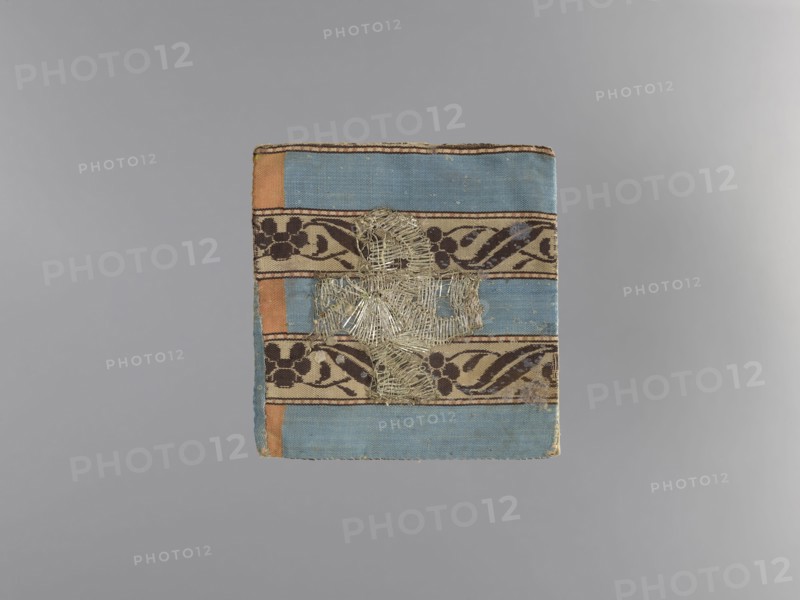 18th century; liturgical parament church parament with flowers and plant ornaments; sacred art; churches and chapels furnishings; height 13 cm, width 13 cm., A rectangular textile with horizontal bands in blue and tan, accented by vertical orange strips at the sides, featuring decorative dark brown floral and vine patterns on the tan bands, with a central area of frayed metallic threads forming an irregular cross-like shape; the textile shows worn edges and small areas of discoloration against a neutral gray background.