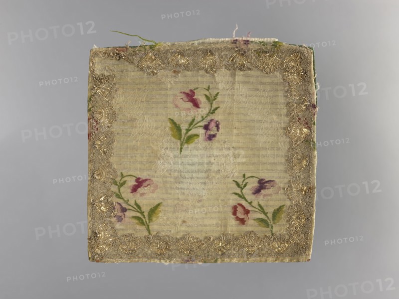 church parament; church paraments; flowers, leaves, plant ornaments; sacred art; churches and chapels furnishings; 2nd half of the 18th century; France; liturgical parament; height 19 cm; width 19 cm, A square piece of fabric with a central panel of light beige or cream-colored woven material showing faint horizontal texture, bordered by an ornate trim with metallic gold threads; within the central area a group of small floral motifs rendered in shades of pink, purple, red, and green with leafy stems; the backing and edges show hints of green and muted red, and the background behind the fabric is a smooth gray.