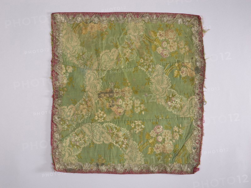 Velum; creator, church paraments; liturgical parament; flowers, leaves, plant ornaments; sacred art; churches and chapels furnishings; 2nd half of the 18th century; France; height 49 cm; width 45 cm; repossession provenance, A rectangular piece of fabric with a green background patterned with floral and paisley-like designs in pale cream, soft pink, and muted yellow, bordered by a narrow strip of faded red fabric and an ornate trim in metallic silver; the fabric shows areas of wear and small darkened spots, with a few creases and slight fraying along the edges, all set against a pale neutral background.