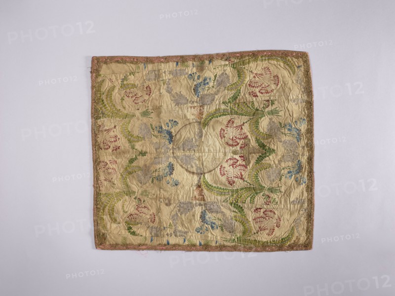 Velum;, liturgical parament church paraments; 18th century, France; sacred art; churches and chapels furnishings; flowers, leaves, plant ornaments; height 42.5 cm, width 48.5 cm., A square textile with a pale beige background laid flat on a light gray surface, showing an ornate embroidered pattern with a few curved leafy motifs in green, clusters of floral shapes in red and blue, and some pale gray embroidered elements; the fabric appears slightly wrinkled and worn, with a narrow decorative border in muted pink and gold tones around the edges.