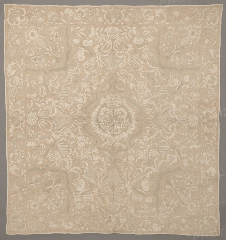 Cope, repossession provenance; clothing accessory; Henryków German: Heinrichau; In the cartouche there are two shields with the monograms CH and DL. Around it are flowers, leaves, and cartouches winding on long stems. Around the edges is a border with a floral tendril motif. Early 18th century, Saxony; fabrics; height 103 cm, width 96 cm, The image shows a large rectangular textile with an overall beige background and intricate off-white embroidered patterns; the design features a central circular medallion with ornate scrolls and laurel-like motifs, surrounded by a symmetrical arrangement of floral and foliate elements, curved vine-like shapes, shield-like forms, and scalloped panels; a decorative border runs along the edges with repeating floral motifs and small ornamental panels; the embroidery creates subtle raised texture and tonal contrast against the beige ground, and the colors visible are beige and off-white.