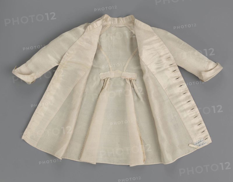Children's dress made of cream satin. Long, fitted at the top, slightly flared at the bottom. Fastened at the front to the neck with 12 small, covered buttons. Low, stand-up collar. Long, set-in sleeves, finished with turn-up cuffs. Widened at the back, from waist level down, by gathered pleats. Unaltered. 2nd half of the 18th century, Poland. Garment; length 49 cm, width 100 cm bottom., A light-colored, open coat or dress laid flat on a gray background, showing the interior with a gathered skirt section and a small decorative tab at the waist; the garment has long sleeves with turned cuffs and visible seam lines and buttonholes along one front edge; the fabric appears smooth and slightly sheer in places, with subtle variations in shading; visible colors include cream, off-white, pale beige, and gray from the background.