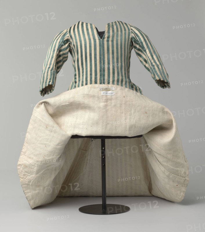 Children's dress, 18th/19th century, Silesia. Children's dress made of satin with green and cream stripes. Fitted at the top, flared at the bottom, extended at the back by a semicircular train. The bodice is slit at the back and laced. The neckline is semicircular with a sharp V-neck at the center front. Long sleeves, cut in an arc at elbow height, finished with small close-fitting cuffs fastened with covered buttons. Lining made of white linen canvas. Unaltered. Length 49 cm. Classification: clothing, children's clothing; fashion; provenance: repossession., A striped garment with vertical teal and cream stripes on the bodice and sleeves sits on a stand, showing puffy sleeves and a V-shaped neckline; a large cream-colored skirt or underskirt with a textured woven surface is folded outward at the waist, revealing the striped interior lining and a darker support structure beneath; the visible colors are teal, cream, off-white, dark gray, and black, and the background is a smooth light gr...