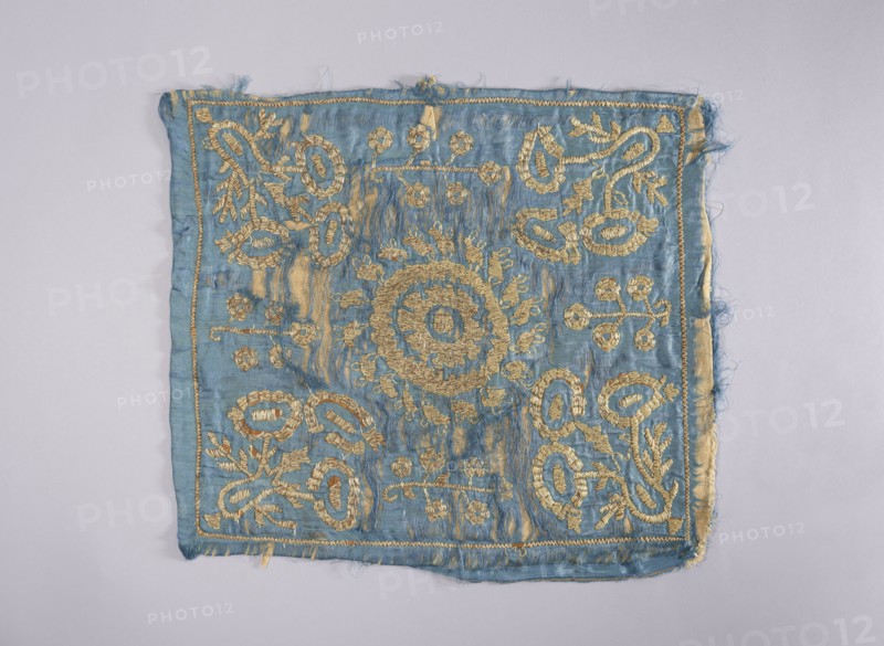 Embroidered vellum, church paraments; flowers, leaves, plant ornaments; sacred art; churches and chapels furnishings. Velum made of blue silk satin, embroidered with silver thread. In the center, a rosette consisting of two ovals. In the corners, large, stylized, schematically treated bouquets of flowers, between which very simplified, smaller flower trees. For reinforcement, embroidered on beige woolen fabric. Lining detached. 2nd half of the 17th century, Poland; liturgical parament; height 52 cm, width 44 cm., A square piece of fabric with frayed edges laid flat on a pale background, predominantly light blue with metallic gold embroidery forming a central circular motif and repeating floral and looping decorative patterns toward the corners and edges; the blue cloth shows areas of wear and slight discoloration where the gold thread is denser, and the embroidery thread has a textured, raised appearance contrasting with the smoother fabric.