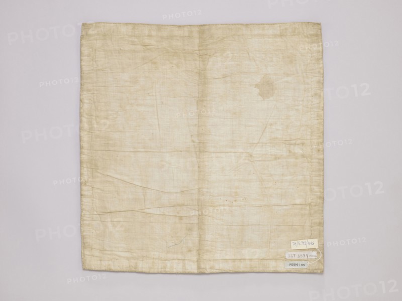 Vellum, vellum church parament with floral ornaments, satin vellum with floral ornament, late 19th century, Russia, Germany, liturgical parament, height 43 cm, width 42 cm, A rectangular piece of fabric lies flat against a neutral background, showing a faded, off-white to beige color with visible creases and folds across its surface; the fabric has slightly darker beige stitching along the edges and a few small irregular discoloration spots and a larger, irregular stain toward the upper right area; there is a group of small pale labels attached near the lower right corner, and the background appears light gray.