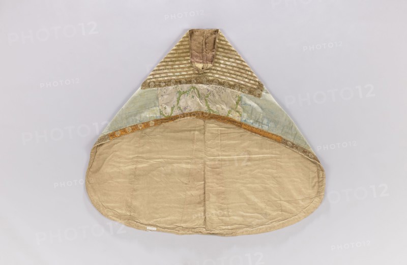 Church cope, unknown origin, decorated with flowers and leaves, plant ornaments; liturgical vestment for church use; sacred art; used in churches and chapels furnishings; 18th century, France; height 133 cm, width 149 cm, An item with a triangular top and rounded lower edge lays flat against a pale background; the upper triangular section has striped fabric in muted gold and cream, flanked by textured gold trim, below which are panels of faded green, blue, and beige fabric with subtle floral and vine patterns, separated from the lower section by a narrow band of orange-gold trim; the large rounded lower area is made of plain beige fabric with visible seams and slight creases, and the overall color palette includes beige, gold, cream, green, blue, and orange-gold.