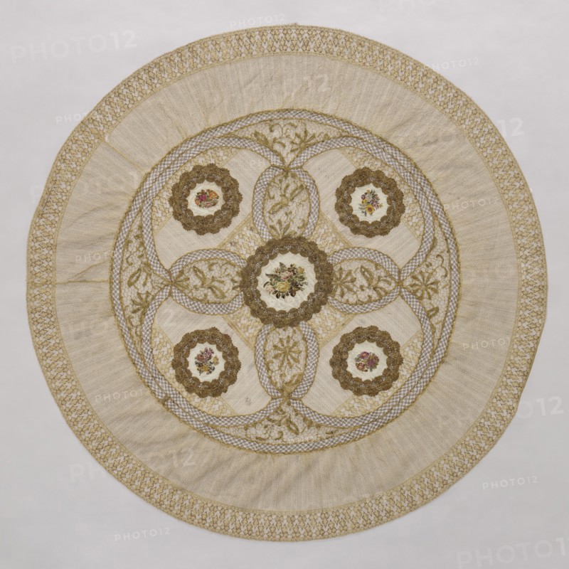 Doily, doilies, decorative fabric, lace, embroidery. Round doily sewn from two pieces of machine lace, bobbin lace, and fabric with colorful petit point embroidery. Five medallions are arranged in the central circle, each surrounded by bobbin lace, with four oval lace medallions with floral branches between the medallions. Mid-19th century, Europe. Fabric, lace; diameter 115 cm., A circular textile with a central round medallion and a symmetrical arrangement of smaller round motifs, all set within concentric ornamental bands; the piece features intricate lacework and embroidered floral designs, with scalloped floral frames around the round motifs, interlaced ribbon-like patterns connecting them, and leaf-and-vine motifs filling oval spaces; the colors visible are cream, beige, gold, brown, green, pink, and red; the outermost edge shows a decorative woven border, followed by a wide pleated or gathered band, then a fine netted area leading into the central embellished section.
