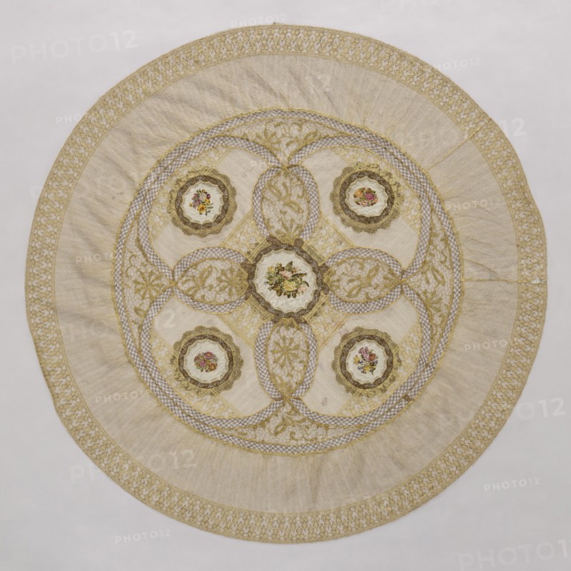 Doily, unknown creator; round doily sewn from two pieces of machine lace, bobbin lace, and fabric with colorful petit point embroidery. Five medallions arranged in the central circle, each surrounded by bobbin lace, with four oval lace medallions with floral branches between the medallions. Mid-19th century, Europe. Fabric, lace. Diameter 115 cm., A circular textile with a central round medallion surrounded by a decorative ring and an outer border, featuring a pale beige main background with gold embroidery and trim, a few scalloped circular appliqués each containing small floral motifs in pink, red, yellow, and green, several interlacing gold embroidered bands forming petal-like shapes, and areas of cream and off-white fabric textures; the outer rim shows a patterned gold lace-like band against the beige, and the overall piece displays a mix of subtle beige, cream, gold, pink, red, yellow, and green colors.