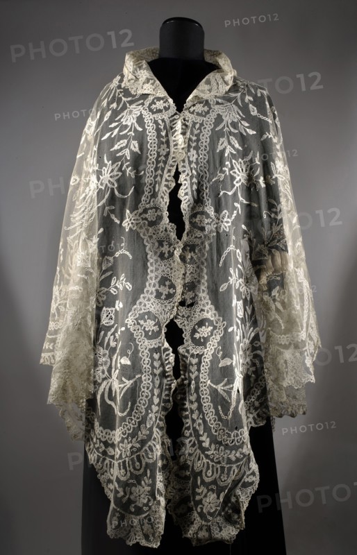 Cape, plant ornaments, floral ornaments, fashion, women's fashion, fashion accessories, capes, lace. Machine-made tulle background with a floral tendril pattern sewn from hand-made bobbin and partly needle-sewn lace. In the center is a motif of a small flower tree, surrounded by flower sprigs. 2nd half of the 19th century, Brussels. Clothing accessory, lace. Height 248 cm, width 123 cm., A sheer lace garment displayed on a black mannequin, featuring intricate floral and scrollwork embroidery in off-white and cream tones on a translucent net background, with a high collar and scalloped edges; the lace shows layered panels and flowing sleeves with delicate trim, and the backdrop is a neutral gray.