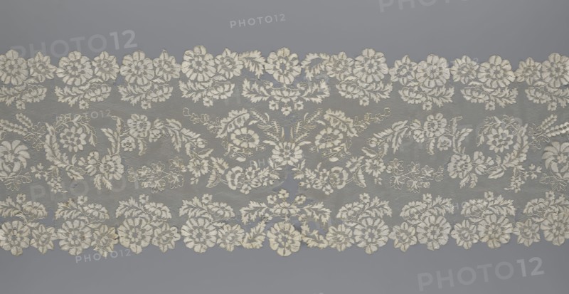 Shawl, shawl; purchase provenance; clothing accessory; women's clothing; fashion; lace with serrated edges; design includes twigs with two flowers in the bends and curling flower twigs in the middle part; 19th century, Europe; length 48 cm, width 260 cm., A wide horizontal piece of lace with a repeated floral pattern in off-white and cream tones set against a muted gray background, featuring a scalloped top and bottom edge formed by clusters of embroidered flowers, leaves, and curling vines, with several larger rosette-like blossoms and a group of smaller filler flowers and foliage arranged symmetrically across the fabric, the lace showing variations in texture where the dense embroidery contrasts with the finer netted sections, and a few tiny irregularities in the thread color and weave.