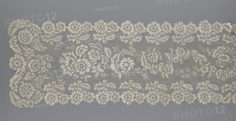 Shawl, 19th century, Europe. Lace shawl, women's clothing accessory; serrated edges; design: in the bends twigs with two flowers, middle part with curling flower twigs. Length 48 cm, width 260 cm., A wide piece of lace fabric laid flat, showing an intricate floral pattern with scalloped edges; the lace features a group of large and small flower motifs, leafy tendrils, and swirling vine shapes arranged in horizontal bands with repeating clusters; the background netting is fine and sheer, while the embroidered elements are raised and textured; visible colors include off-white or cream for the lace and a muted gray background.