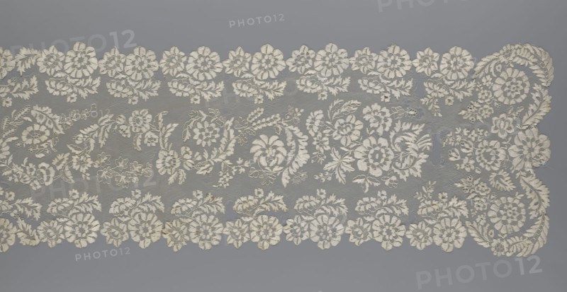 Shawl, shawl; clothing accessory; women's clothing; fashion; lace with serrated edges; design features: twigs with two flowers in the bends and curling flower twigs in the middle part; 19th century, Europe; length 48 cm, width 260 cm., The image shows a length of intricately patterned lace laid flat against a smooth background. The lace features a repeating floral motif with a scalloped edge composed of round flower shapes and leafy scrolls; between the scallops are flowing garlands of flowers and foliage woven into a fine net ground. A few larger floral clusters and stylized leaves form a central band, while several smaller blossoms and sprigs fill the spaces, creating an overall dense, delicate pattern. Visible colors include off-white for the lace and a cool gray for the background.