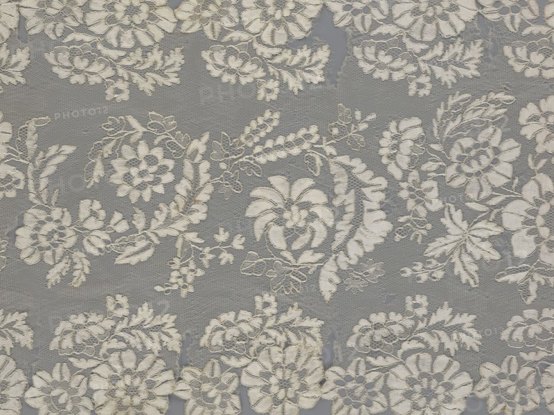 Shawl, shawl, clothing accessory, women's clothing, fashion, lace, serrated edges, in their bends twigs with two flowers, in the middle part curling flower twigs, 19th century, Europe, lace, length 48 cm, width 260 cm, A patterned textile with a repeating floral lace design in shades of off-white and beige set against a muted gray background, showing a group of embroidered flowers, leaves, and vine motifs interconnected across a fine netted mesh, with several circular and petal-shaped flower heads, feathery leaves, small clustered buds, and scalloped edges; the stitching creates subtle raised textures and shadows, and there are areas where the netting shows slight irregularities.