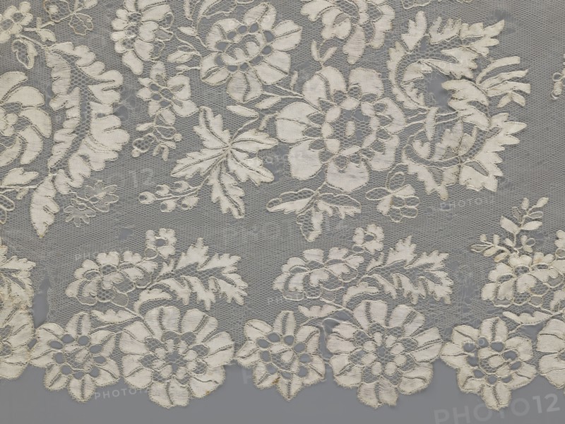 Shawl, shawl; 19th century, Europe; clothing accessory for women; lace with serrated edges; decorative design: in the bends twigs with two flowers, in the middle part curling flower twigs; dimensions: length 48 cm, width 260 cm., A piece of lace fabric with an intricate floral pattern in shades of off-white and cream on a gray background, showing a mesh-like net base with embroidered flowers, leaves, and scroll motifs, scalloped edges along one side, and areas where the lace overlays the background creating subtle shadows and varying texture.