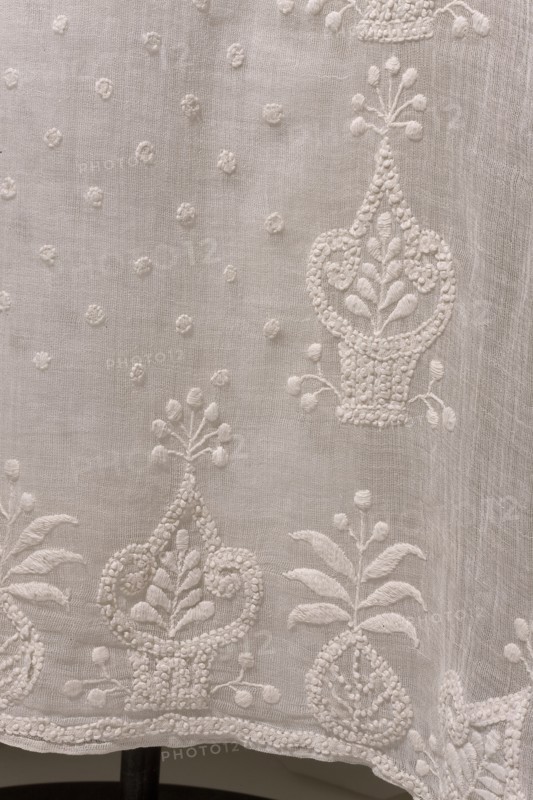 Dress with train, Dress of white muslin, early 19th century, Silesia. High waist, with a long train; deep-cut square neckline, cinched at the front with a drawstring. Three-quarter set-in sleeves, gathered at the top and flared at the bottom, with embroidered inserts featuring smaller geometric patterns. Skirt sewn to the bodice, narrow at the front and fuller at the back, long train. Front decorated with embroidery: between two rows of stylized flower baskets are small scatterings of polka dots. Hem and train decorated with stylized flower baskets. Empire style. Trains clothing, geometric ornaments, basket with flowers ornament. Length 131 cm front, 230 cm back; hem circumference 408 cm. Associated with Ludwika Queen of Prussia; 1776–1810 — souvenirs; repossession, A close-up view of a textured light-colored fabric with embroidered floral and botanical motifs in the same pale color, showing several ornate vase-like shapes, leaves, small round raised dots, and delicate stitched borders...