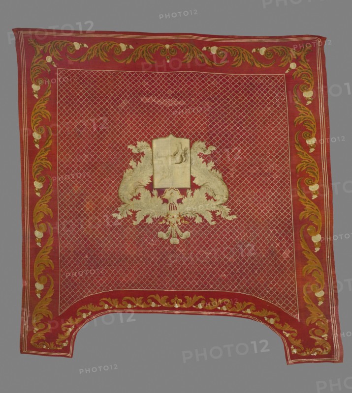 Saddle pad — kilim in a near-square form with slightly elongated upper corners. Main field: rhomboidal mesh ornament with small meshes; central rectangular shield depicting the Boncza unicorn coat of arms, framed at the top by wide and long labres. Surrounded by a border with acanthus leaf motifs arranged in a tendril. Colors: dark red background; ornament in cream, celadon, and tobacco. Date: 1830s. Place of origin: Poland. Material: fabrics. Dimensions: height 248 cm, width 235 cm., A rectangular textile with a dominant red field and a patterned lattice of lighter lines filling the central area, bordered by a wide red band decorated with scrolling floral and acanthus motifs in shades of gold, ochre, and pale cream, with the border motifs repeated along all sides; in the center sits an ornate symmetrical emblem formed of pale cream and gold embroidered foliage and scrollwork surrounding a plain pale rectangular panel, and the lower edge of the textile has a curved cutout with matching...