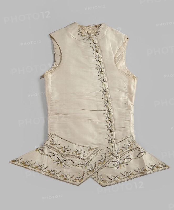Vest, plant motifs, garlands ornament; men's clothing, 18th century, embroidery, waistcoat. Men's waistcoat of cream silk, embroidered. Straight cut, without a collar; trapezoidally cut hems; low-set pockets covered with flaps. Fastened to the neck with 12 covered and embroidered buttons; identical buttons under the pocket flaps. Embroidery decorates the edges at the closure, the pocket flaps, and the flaps under the pockets. Small floral garlands and tied bundles of grass. Embroidery with satin stitch. Predominant colors: yellow, green, and dark purple. Laced back, silk lining. Fabric-covered and embroidered buttons. 1780s, Europe; Silesia. Garment; length 70 cm., A sleeveless, light beige silk garment laid flat against a pale gray background, showing a high neckline and an asymmetrical front opening with a row of covered buttons; the fabric has a subtle sheen with visible creases and seams, and ornate floral embroidery along the front opening and hem in shades of green, gold, brown, ...