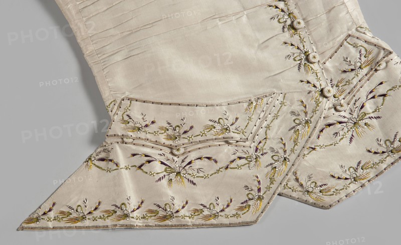 Men's waistcoat of cream silk, embroidered. Straight cut, without a collar; trapezoidally cut hems; low-set pockets covered with flaps. Fastened to the neck with 12 covered and embroidered buttons; identical buttons under the pocket flaps. Embroidery decorates the edges at the closure, the pocket flaps, and the flaps under the pockets. Small floral garlands and tied bundles of grass. Embroidery with satin stitch. Predominant colors: yellow, green, and dark purple. Laced back, silk lining. Fabric-covered and embroidered buttons. 1780s, Silesia Europe. Length 70 cm., plant motifs; garlands; clothing; men's clothing; waistcoat; garment; repossession provenance., The image shows a section of a pale cream-colored garment with a smooth, slightly shiny fabric and decorative embroidery. There is a pointed flap and a folded edge with small rounded covered buttons along a seam. The embroidery forms repeating floral and leafy motifs in muted yellow, olive green, deep purple, and a pale off-white,...