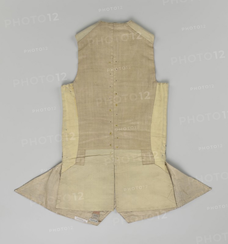 Vest, Floral and plant ornaments on brocaded silk fabric. Predominant ornamental colors: silver, plum, green, and raspberry. Front with motifs arranged around the edges; fastened with 12 buttons embroidered with gold threads; no collar. Side flaps with flapped pockets, three buttons under the flaps as for the closure. Back: bottom and sides of fabric as on the front, cut-out pieces plain without brocade. Cream silk lining. 1740–1760, England fabric possibly from Silesia. Garment; length 74 cm., A sleeveless tailored garment laid flat, showing the back with a fitted waist and two flared skirt-like panels at the bottom; the fabric appears in shades of beige, cream, and light tan with visible stitching lines, seam edges, and small reinforcement patches; there are a few small puncture-like markings along the center back seam and several subtle creases and worn areas on the skirt panels; the shoulders have slightly darker beige patches and the interior lining visible at the bottom edge is a...