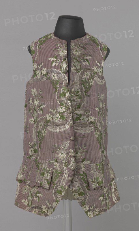 Vest, floral ornaments, patterned fabrics, Henryków German: Heinrichau - museum repository, fastened to the neck with 11 buttons. Flaps on the sides, below them 4 buttons as for the fastening, embroidered in gold. The back is made of brown canvas at the top and fabric at the bottom as on the front. 1750s, Silesia, garment, length 76 cm, A sleeveless, knee-length garment displayed on a black mannequin torso, made of a ribbed fabric in a muted mauve color with an all-over floral brocade in shades of green and off-white; the front has a narrow standing neckline and a vertical row of fabric-covered buttons and decorative loops, with a short pleated peplum and flap pockets near the hem, and the floral pattern forms scalloped borders and clusters of leaves and blossoms against the mauve ground.