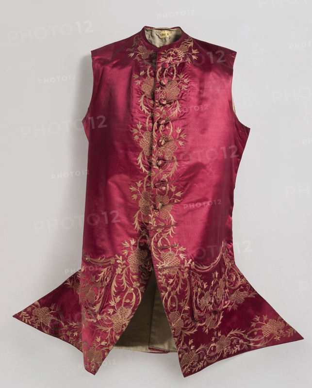 Men's vest made of cherry satin, embroidered. Straight cut, without sleeves or collar. Diagonal hemline, side pockets covered with large flaps, three covered buttons under each flap. Fastened to the neck with 11 covered and embroidered buttons. Circa 1740–1770, Silesia. Length 78 cm., repossession, Floral ornaments., A sleeveless garment displayed against a pale background, made of a shiny deep red fabric with a vertical panel of intricate gold embroidery running down the center and expanding into ornate floral and leafy patterns toward the lower flared sections, featuring decorative fabric-covered buttons along the front panel, a rounded high collar with matching embroidery, and a contrasting lighter-colored inner lining visible where the front splits; visible colors include deep red, gold, a light beige inner lining, and the pale neutral background.