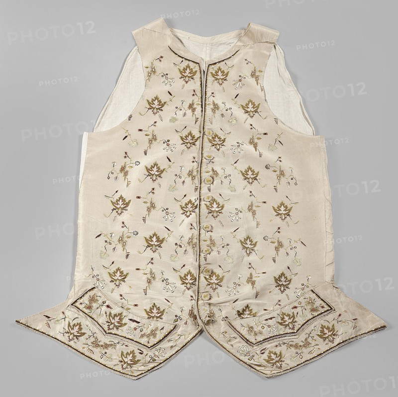 Vest, 2nd half of the 18th century, Poland. Garment, length 74 cm. Fastened to the neck with 9 covered and embroidered buttons. Side flaps with pockets underneath. Laced canvas back. Floral and geometric ornaments., A sleeveless garment laid flat on a neutral gray background, made of a pale cream fabric with a slightly shiny finish and a lighter cream inner lining visible at the arm openings; the front panels feature a repeated floral embroidery pattern in shades of olive green, muted gold, soft brown, and pale beige with small accents of darker brown and subtle touches of light green, and a vertical trim runs down the center front; the hem has two pointed flaps with additional embroidered floral motifs and decorative borders in the same color palette, and the garment shows stitched seams and a small standing collar in the same pale cream tone.