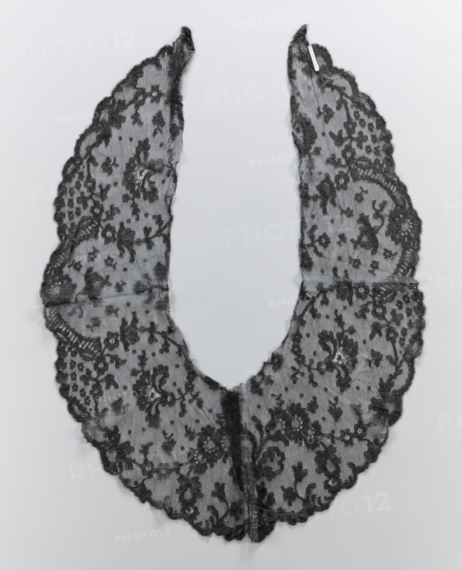 Collar, lace clothing accessory for women's fashion. Around the outer serrated edge are shell-and-lace motifs. The collar is widest in the middle, tapering at the elongated ends. The inner edges are cut and unsecured. 2nd half of the 19th century. Probable origin: Belgium or France. Materials: lace; dimensions: width 23 cm, length 81 cm., A delicate scalloped lace collar in black is laid flat on a pale gray background, forming a U-shape with two elongated curved panels meeting at the bottom; the lace features an ornate floral pattern with clusters of flowers, leaves, and scrolling vines worked into a fine mesh, and the edges are finished with a scalloped trim.