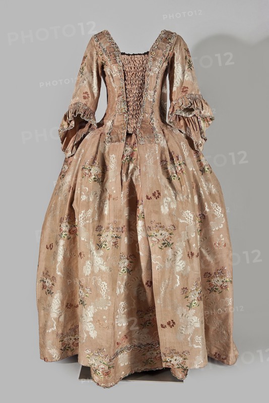 Two-piece robe à la française dress, silk in the colour of dried rose with motifs of colourful flower bouquets and winding white lace ribbons. Overskirt manteau open at the front, with a triple pleat extending almost the entire back and forming a train. Bodice on linen lace with boning, laced at the front; deep square neckline. Narrow elbow-length set-in sleeves trimmed with ruffles of the same fabric. Neckline and bodice edges trimmed with a pleated ruffle decorated with multicolored cord trimmings twisted into tiny flowers. No lining. Skirt gathered with symmetrical pleats, spread to the sides. Patterned fabric; fabric report: height 24.5 cm, width 48.5 cm; width of fabric piece 54 cm. Dated 1760–1765. Origin/attribution: Silesia; possibly France. Length 158 cm. Features: floral ornaments, ribbon ornament, flower bouquet motifs, ruffles; women's 18th-century clothing., A full-length historic-style dress displayed against a plain background, made of a shiny pale pinkish-beige fabric p...