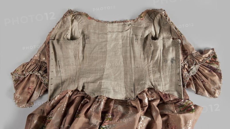 Two-piece robe à la française dress, silk in the colour of dried rose with motifs of colourful flower bouquets and winding white lace ribbons. Overskirt manteau open at the front, with a triple pleat extending almost the full width of the back and forming a train. Bodice on linen lace with boning, laced at the front; deep square neckline. Narrow elbow-length set-in sleeves trimmed with ruffles of the same fabric. Neckline and bodice edges trimmed with a pleated ruffle decorated with multicolored cord trimmings twisted into tiny flowers. No lining. Skirt gathered with symmetrical pleats and spread to the sides. Patterned fabric with floral ornaments, ribbon ornaments and flower-bouquet motifs. Ruffles and other clothing decoration. Fabric report: height 24.5 cm, width 48.5 cm; width of fabric piece 54 cm. Date: 1760–1765. Place of use: Silesia; probable origin: France. Length: 158 cm., Women's fashion, 18th century., A worn garment laid flat showing an inner lining of coarse beige fabri...