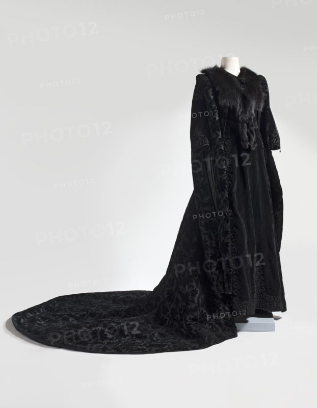 Dress, Art Nouveau style, late 19th century, Poland, attributed, Women's garment, lightly fitted at the back waist with a small train tail in the form of a minimized bustle. Shallow triangular neckline. Sleeves of uncertain length, slightly raised at the cape, cut in two pieces and ending with six pendants with black rhinestone balls. Fitted back with a 23-hook fastening. Embroidery decorates the edges of the neckline, the caps and bottoms of the sleeves, the cutting lines, and the lower hem of the skirt. Small flat pocket sewn on the left side below the waist. Materials/style details: trains garment, meander ornament. Dimensions: length 163 cm back, circumference 264 cm bottom., A full-length black velvet cloak with a long sweeping train is displayed on a headless mannequin; the cloak has a dark fur collar and fur trim along the front opening, ornate raised patterns in the fabric creating floral and geometric motifs, long wide sleeves with decorative detailing, and the train spreads a...