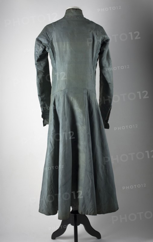 Zupan made of steel-blue grosgrain, long, fastened at the front with leatherwork buttons. Fitted at the top, slightly flared at the bottom. High, stand-up collar. Long, set-in sleeves, finished with turn-up cuffs fastened with hooks. Side pockets. The back is cut with a pole like that of a kontusz, widened from the waist by semi-flared pleats. Lining made of gray linen and indigo-dyed linen, impregnated. Hand-sewn, 18th century, Poland; men's clothing, national costume, length 122 cm, circumference 182 cm bottom. Polish clothing; materials: silk, steel-blue grosgrain, gray and dyed canvas. Creator: unknown Czechman., A long sleeved dress displayed on a mannequin stand, shown from the back, in shades of bluish-green and gray with visible creases and seams; the bodice is fitted with a high collar and long sleeves that taper at the cuffs, and the skirt flares into a flowing, pleated shape reaching toward the floor; the mannequin stand is black and supports the garment against a neutral gr...