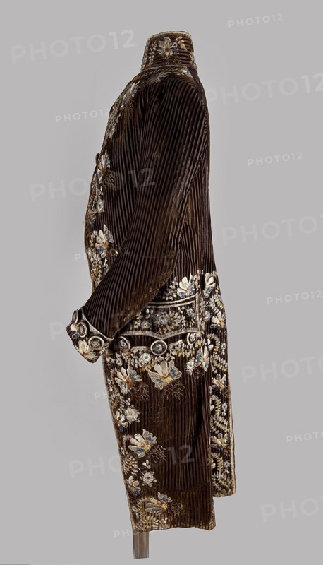 Habit à la française, tailcoat, floral ornaments, clothing, men's clothing, men's fashion, 18th century clothing. Habit of striped brown velvet, embroidered with fanciful flowers with large stamens and ears of grass. Fitted at the top, widened by pleats at the bottom, at the back, from the waist down. Long, set-in sleeves, cut in an arc at elbow height, finished with cuffs. Fastened with buttons; the skirts are cut in an arc, overlapping at the chest. Embroidery along the edges of the pockets, on the cuffs, pocket flaps, collar, and back pleats. The back is cut with a center seam; the back pleats do not overlap. Three buttons under the pocket flaps and on the cuffs. 1780s, Silesia, garment, length 111 cm., A side view of a long, dark brown velvet coat with vertical ribbed texture, decorated with embroidered floral motifs in colors including white, cream, blue, and gold; the coat has a high collar and long sleeves, ornate cuffs, and a waist area embellished with circular decorative elem...