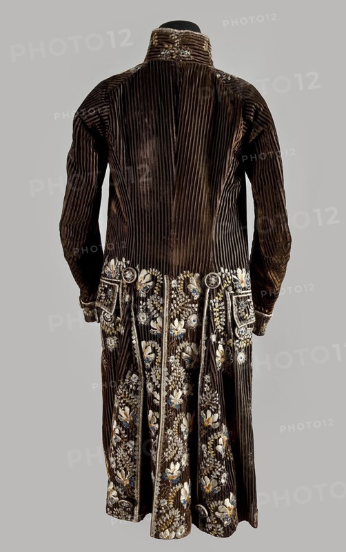 Habit à la française, tailcoat, floral ornaments, clothing, men's clothing, men's fashion, 18th century clothing. Habit of striped brown velvet, embroidered with fanciful flowers with large stamens and ears of grass. Fitted at the top, widened by pleats at the bottom, at the back, from the waist down. Long, set-in sleeves, cut in an arc at elbow height, finished with cuffs. Fastened with buttons, the skirts are cut in an arc, overlapping at the chest. Embroidery along the edges of the pockets, on the cuffs, pocket flaps, collar, and back pleats. The back is cut with a center seam; the back pleats do not overlap. Three buttons under the pocket flaps and on the cuffs. 1780s, Silesia, garment, length 111 cm, A long, dark brown velvet coat with vertical ribbing visible across the back and sleeves, shown from the rear on a neutral gray background; the lower portion of the coat is decorated with detailed floral embroidery in colors including white, light blue, gold, and pale yellow, with add...