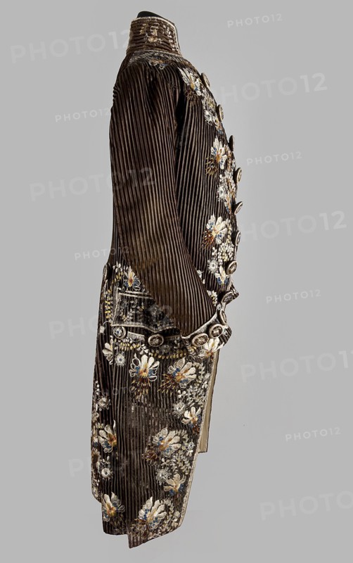 Habit à la française, tailcoat, floral ornaments, clothing, men's clothing, men's fashion, 18th century clothing. Habit of striped brown velvet, embroidered with fanciful flowers with large stamens and ears of grass. Fitted at the top, widened by pleats at the bottom, at the back, from the waist down. Long, set-in sleeves, cut in an arc at elbow height, finished with cuffs. Fastened with buttons, the skirts are cut in an arc, overlapping at the chest. Embroidery along the edges of the pockets, on the cuffs, pocket flaps, collar, and back pleats. The back is cut with a center seam; the back pleats do not overlap. Three buttons under the pocket flaps and on the cuffs. 1780s, Silesia, garment, length 111 cm, A long dark brown, vertically ribbed coat displayed on a mannequin, shown from the side; the coat has a high stand-up collar and a row of decorative round buttons along the front; it is richly embroidered with floral motifs in white, cream, blue, gold, and a hint of red, concentrated ...
