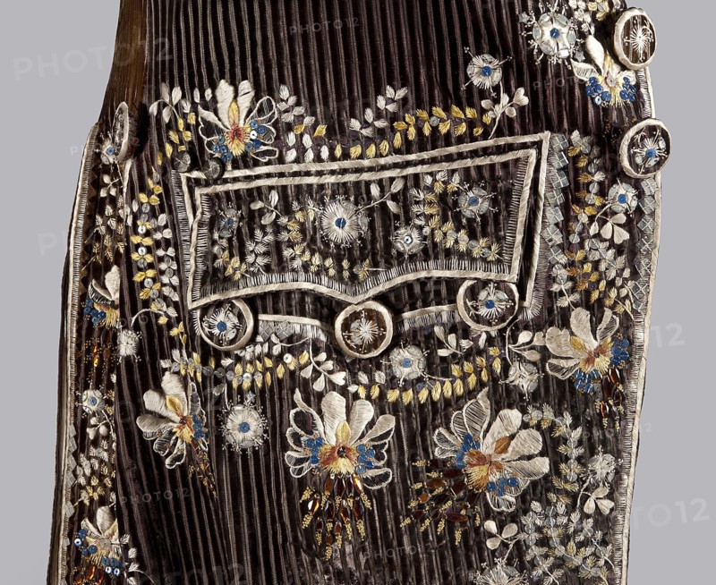 Habit à la française, tailcoat, repossession provenance noted, floral ornaments, men's clothing, men's fashion, 18th century clothing. Habit of striped brown velvet, embroidered with fanciful flowers with large stamens and ears of grass. Fitted at the top, widened by pleats at the bottom at the back from the waist down. Long set-in sleeves, cut in an arc at elbow height, finished with cuffs. Fastened with buttons; the skirts are cut in an arc, overlapping at the chest. Embroidery along the edges of the pockets, on the cuffs, pocket flaps, collar, and back pleats. The back is cut with a center seam; the back pleats do not overlap. Three buttons under the pocket flaps and on the cuffs. 1780s, Silesia, garment, length 111 cm., A close-up of a dark brown ribbed fabric panel with ornate embroidered floral motifs and decorative trim; there is a rectangular flap outlined with light-colored cord and circular button-like decorations, and the surrounding surface shows a group of embroidered flow...